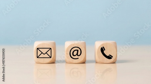 Three wooden cubes with an envelope symbol at sign and telephone icon on a light beige table with a soft blue background in bright studio light showing communication opti