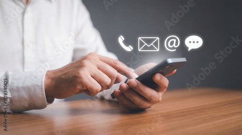 A professionals hands operating a modern smartphone showing glowing digital icons for phone email and message contact on a wooden desk with a dark background