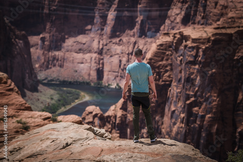 View of a man in a blue shirt gazes out over the precipitous red rock cliffs and the winding river below, bathed in sunlight, Page, Arizona, United States.