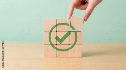 A human hand carefully places a wooden block with a vibrant green checkmark symbol into a grid of blocks on a smooth wooden table against a soft pastel green background d