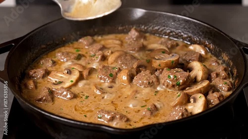 A close-up view of a hand stirring a rich, creamy beef stroganoff with mushrooms in a cast iron skillet, set against a dark kitchen background, showcasing culinary preparation
