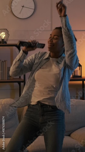 Happy woman singing into a remote control and dancing in her living room at night. Carefree home entertainment and fun. Vertical video.