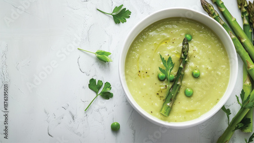 Top-down view of creamy pale green asparagus soup served in a white bowl, garnished with fresh asparagus spears and green herbs, on a clean minimal background
