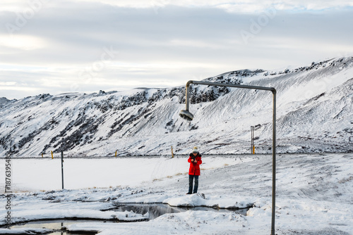View of a person in a red jacket standing near a showerhead in the snow-covered landscape, stark against the pale sky, Myvatn, North Iceland, Iceland.