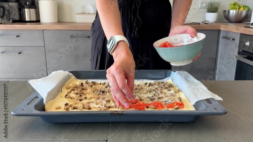Close up of woman hands placing tomato slices on pizza dough on baking tray in home kitchen. Homemade pizza preparation process, cooking routine and everyday food making at home.