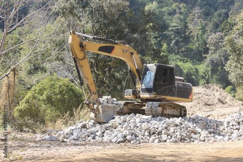 Heavy yellow excavator digging large rock pile with shovel bucket showing industrial strength at sunny outdoor construction site near green forest woods