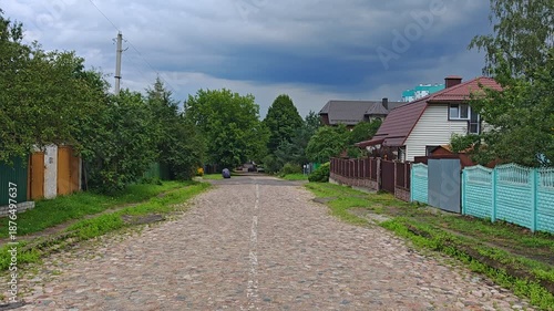 The city street is paved with cobblestones, and the asphalt sidewalks are overgrown with grass. There are trees, fences, houses, parked cars, and power lines along the road. Overcast summer weather