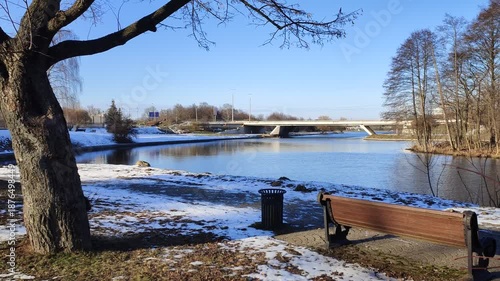 There is a wooden bench on a metal base and an urn on a tiled path near a tree. There is a concrete road bridge over the river and cars are driving. Sunny winter weather and remnants of snow