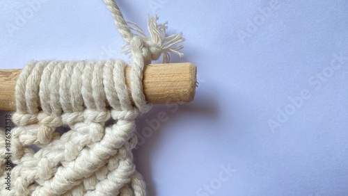 Macrame craft detail of cream colored cotton rope tied to a wooden dowel, showing the texture of handmade knots against a white background.