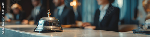 A silver service bell sits on a counter, with people blurred in the background, possibly in a hotel lobby or at a reception desk. 