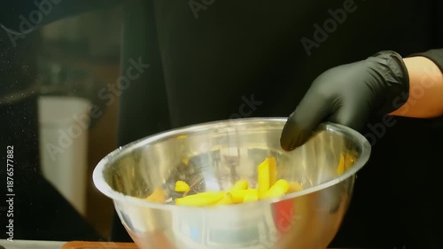 Cook tosses french fries in metal bowl to spread salt. Gloved hand flips bowl upward quickly while fries tumble settling for even seasoning