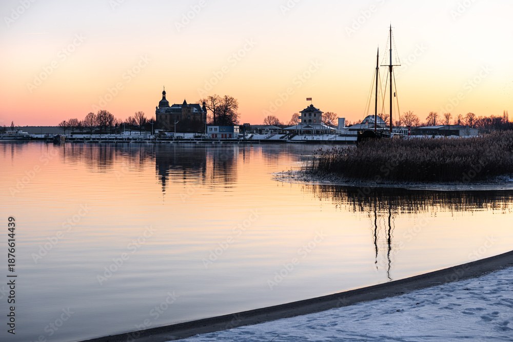 Fototapeta premium Sunset at Goitzsche Lake with view toward the promenade and marina, snow-covered landscape, orange-red sky, winter lakeside scenery in Central Germany.
