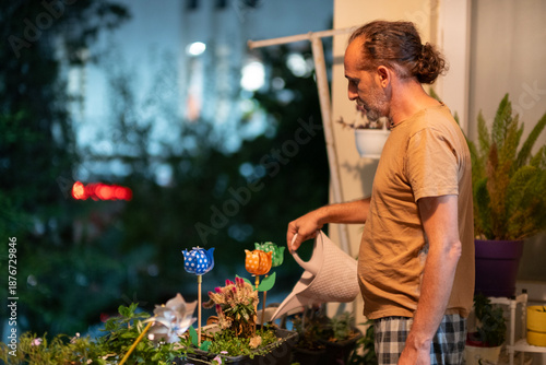 Man Watering Flowers on Balcony. Urban Gardening on Apartment Balcony. Home Gardening and Plant Care Concept. Man Taking Care of Balcony Plants.