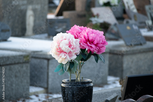 Pink and white artificial peonies in a black vase on a snowy grave in a cemetery