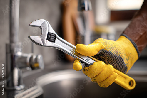 A hand in a yellow glove holds a silver adjustable wrench, ready for plumbing work near a sink in a well-lit room.