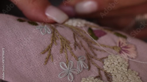 Close-up view of female hands carefully embroidering delicate white flowers onto pink fabric. The detailed needlework features floral patterns and lace edges, showcasing traditional craftsmanship and 