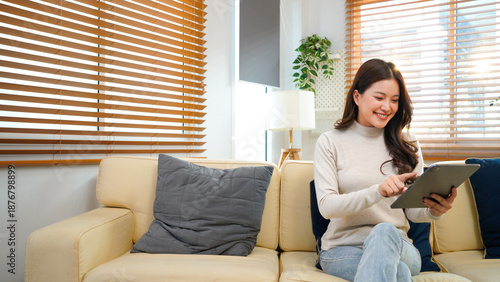 Happy young woman sitting on sofa in modern white living room, using Tablet computer to view content surrounded by bright natural light. Beautifull people and modern technology concept image.