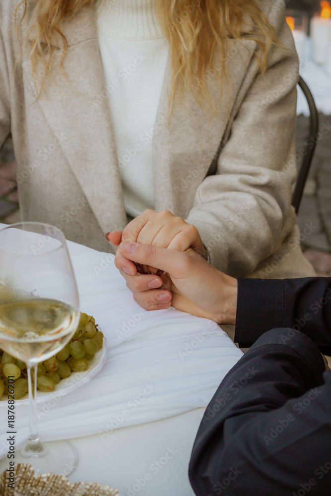 Obraz premium Romantic marriage proposal moment at outdoor cafe with couple holding hands, engagement ring, white wine and grapes. Intimate wedding or engagement scene with elegant table setting and warm atmosphere