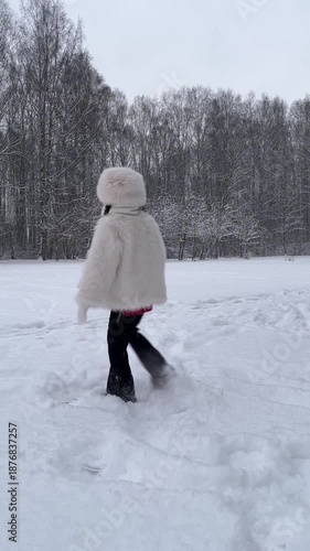 A girl in a white hat and a white fur coat is having fun in a winter snow park