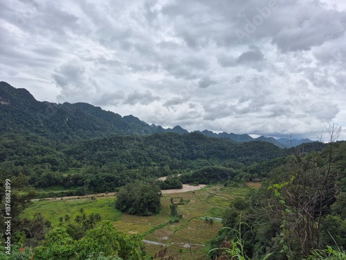 A breathtaking vista reveals lush green mountains and valleys blanketed in dense tropical forest under a dramatic overcast sky.
