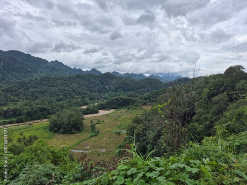 A breathtaking vista reveals lush green mountains and valleys blanketed in dense tropical forest under a dramatic overcast sky.