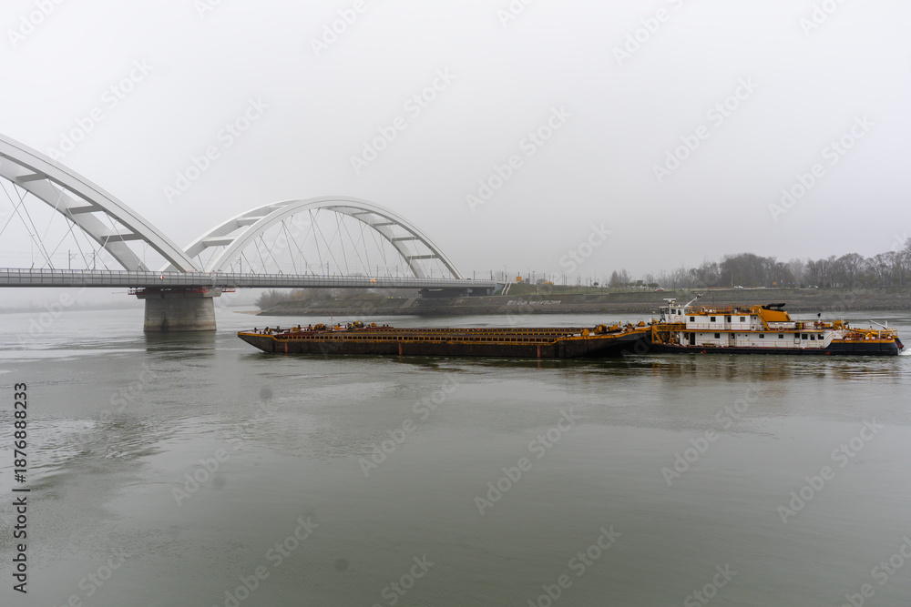 Obraz premium Cargo barge sailing on a wide river beneath a modern arched bridge on a foggy day. Industrial transport, calm water, muted tones, overcast weather, quiet atmosphere
