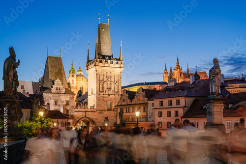 Foto Crowds of people stroll across the historic Charles Bridge in Prague at twilight, with the illuminated Old Town Bridge Tower and Prague Castle standing majestically against a deep blue sky