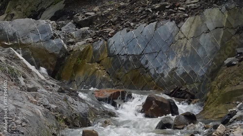 Footage captures a small, fast-flowing mountain stream with white water cascading over dark, jagged rocks. The surrounding rock face features striking, geometric patterns, possibly columnar basalt, wi
