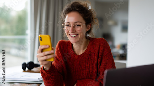 a happy young woman student sitting at a desk with an open laptop, celebrating exciting good news on her smartphone