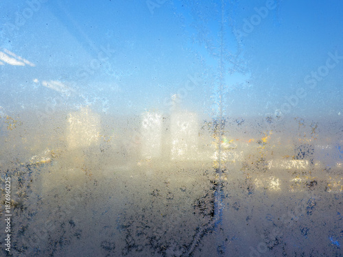 The cold frost on a big window, with a blue sky and some blurry buildings behind it.