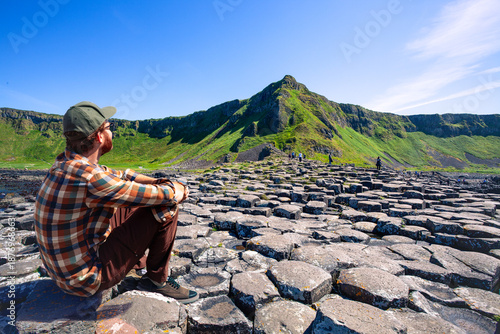 Foto A traveler sits on the unique hexagonal basalt stepping stones of the Giant's Causeway in Northern Ireland, gazing at the dramatic green cliffs under a clear blue sky