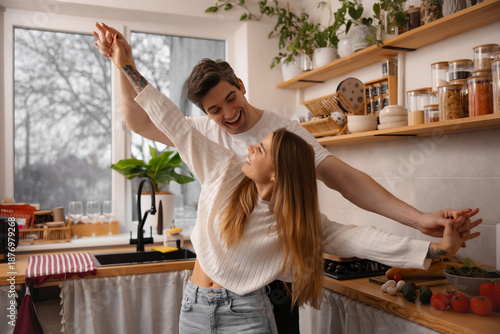 Joyful husband and wife dancing with wide open arms in a bright kitchen