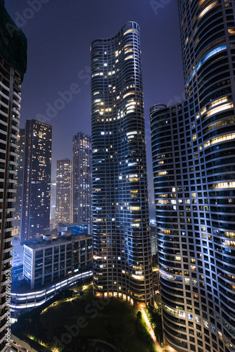 Cuadro en lienzo Dense urban skyline at night with illuminated high-rise buildings emerging throu