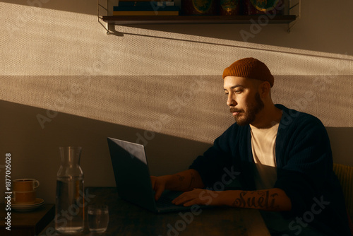 A focused man with a beard and beanie working on his laptop at a wooden table during golden hour. The warm sunset light creates dramatic shadows across the wall in a cozy indoor setting.