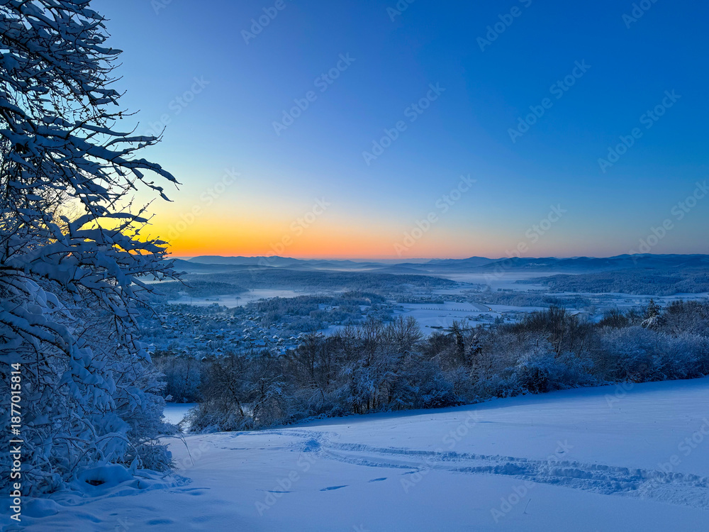 Obraz premium Winter panorama of a snowy valley and distant hills during blue hour at sunrise. Soft light spreads across forests and countryside, blending deep blues with warm tones for a tranquil winter scenery.