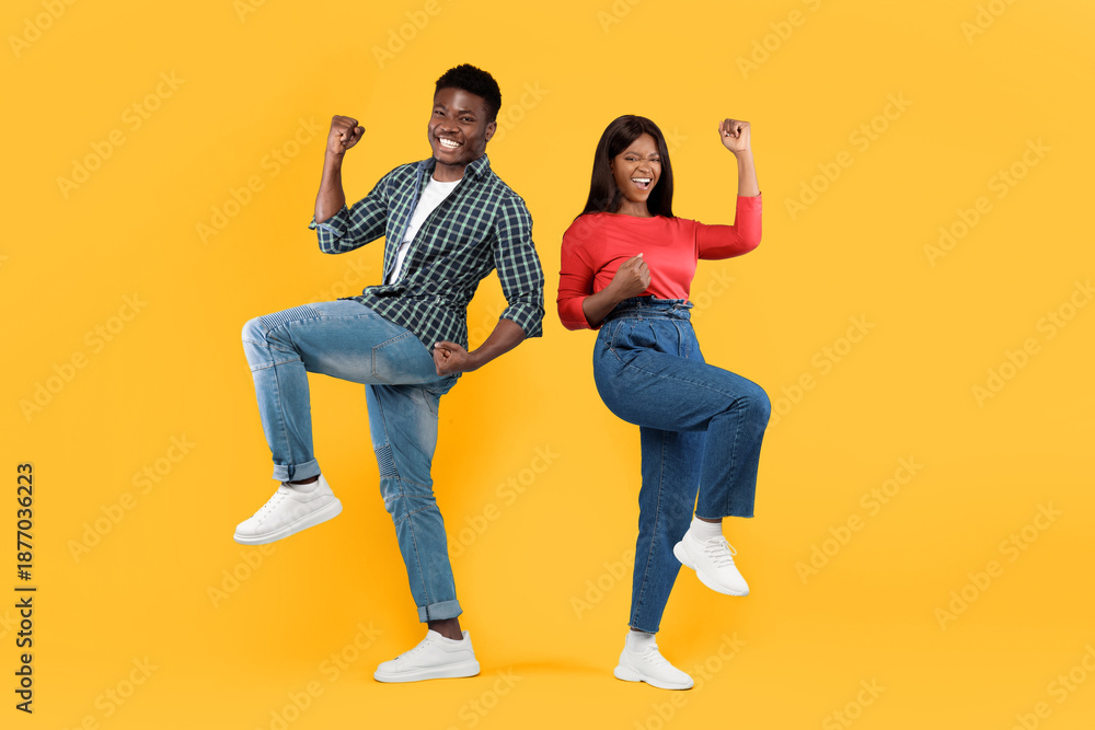 Fototapeta premium Two young adults express happiness by dancing together in playful poses. They wear casual clothes and enjoy the moment against a bright yellow background.