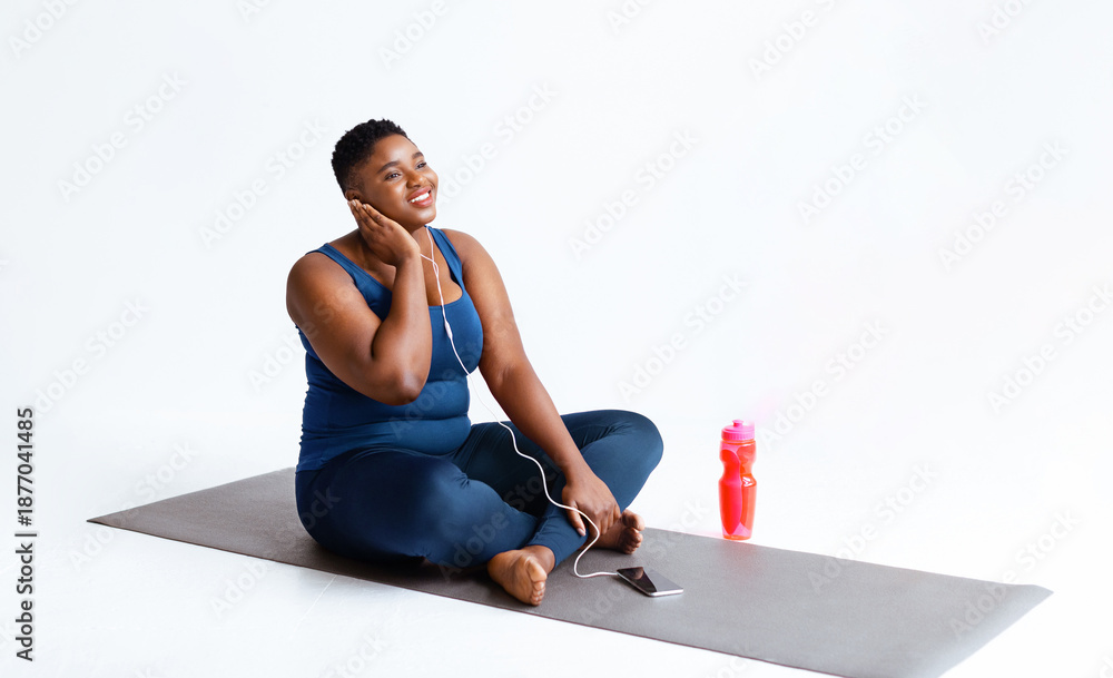 Fototapeta premium A woman sits on a yoga mat while enjoying music through earphones. She looks happy as she relaxes with a water bottle beside her. The setting is simple and bright.