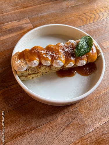 Rolled crepe topped with toasted meringue, caramel sauce and mint leaf, served on a white plate over a wooden table in warm natural light. Amazing dessert in restaurant or cafe