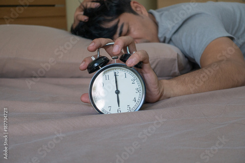 Tired young Asian man sleeping on bed and reaching hand to stop black alarm clock at 6 am. Sleepy male waking up early with headache or hangover in morning bedroom.