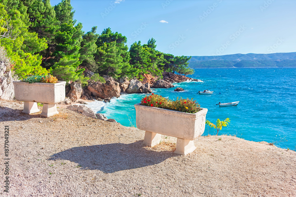 Fototapeta premium Wonderful beach of Croatia. Coastal tranquility. Serene view of Mediterranean Sea, framed by greenery and charming flower boxes. Picturesque coastal landscape with small boats on azure water