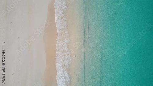 Stunning aerial perspective of a tranquil tropical coastline where soft white sand beautifully merges with the vibrant turquoise ocean, kissed by gentle foamy waves
