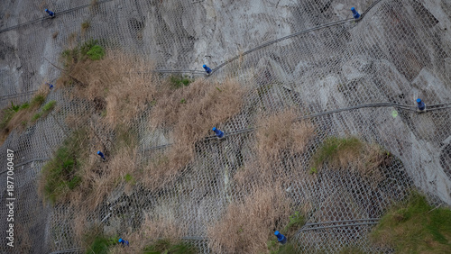 A low-angle detailed view of a steep rocky cliff face reinforced with a heavy-duty steel wire mesh netting designed to prevent rockfalls and landslides.