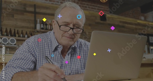 Writing senior man wearing checked shirt, reading glasses, using laptop and noting with pen at cafe