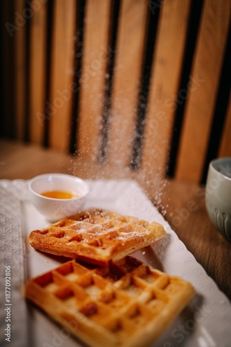 Dynamic food scene of Belgian waffles being sprinkled with powdered sugar. Cozy breakfast mood, warm tones, shallow depth of field and natural light on wooden table