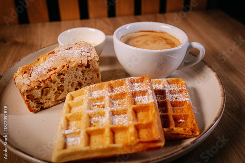 Cozy brunch composition with waffles, homemade cake and coffee served on a ceramic plate. Warm natural light, rustic textures and intimate framing create a welcoming café-style food scene.