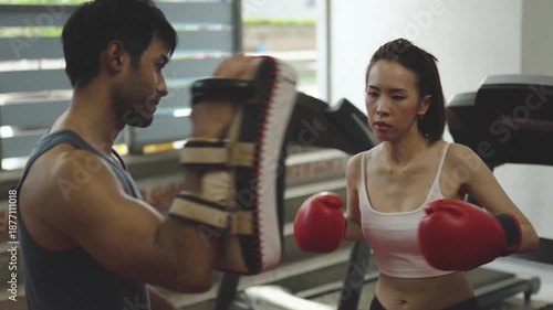 Young woman training boxing with male coach at gym, wearing red gloves and focused expression, promoting strength, fitness, empowerment, and personal discipline.