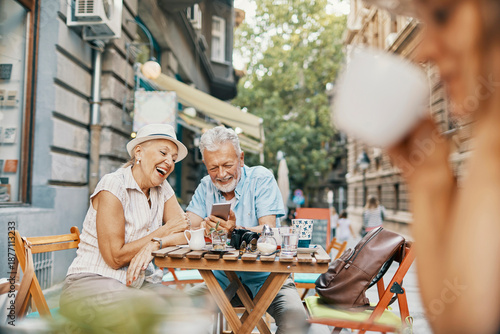 Senior couple laughing over smartphone at city sidewalk cafe