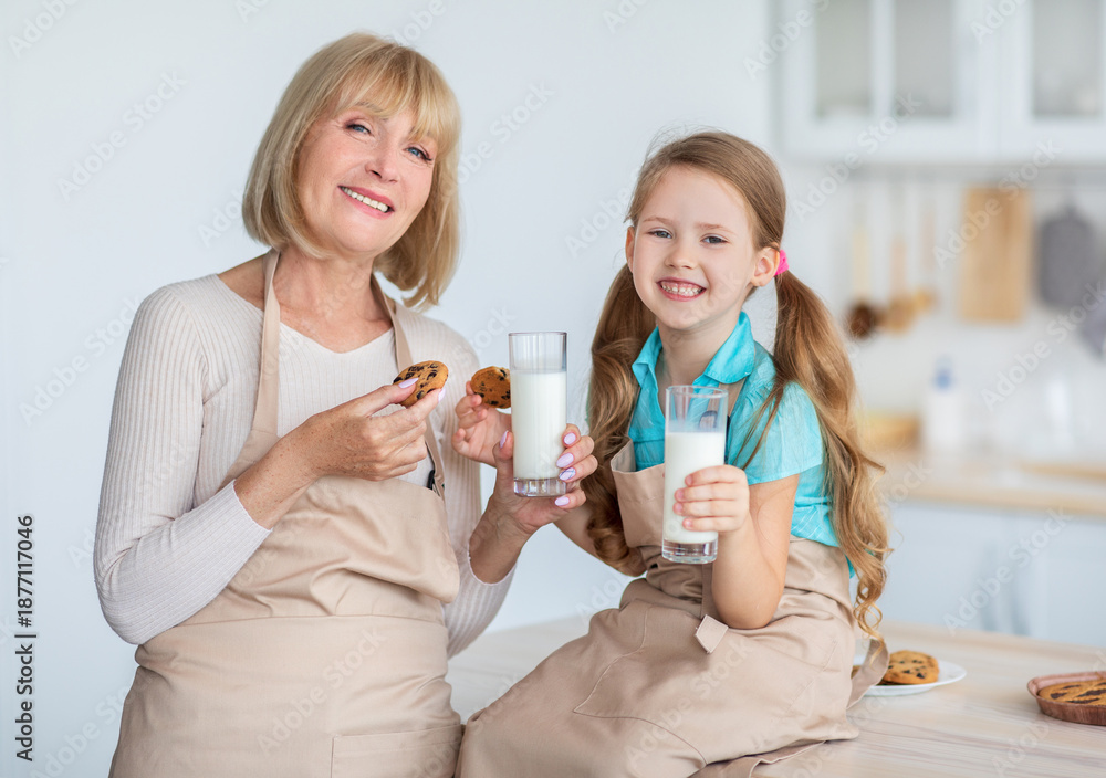 Naklejka premium Snack Time. Portrait of happy granny and little girl enjoying homemade pastry and drinking milk in kitchen, holding fresh baked chocolate chip cookies, smiling and looking at camera, wearing aprons