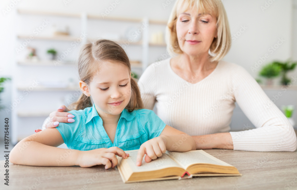 Fototapeta premium Portrait of loving caring mature grandmother teaching cute little girl how to read, sitting at desk with interesting book, spending time together at home in living room. Preschool education concept