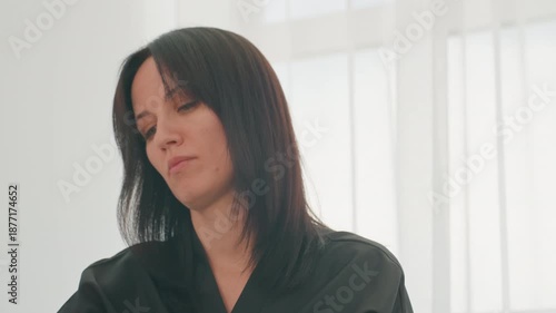 hairstylist examining hair in bright salon, woman in black smock tilting head with calm focused expression, soft natural light through sheer curtains, closeup head and shoulders, stylist assessing